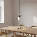 Witte hanglamp met zwart bovenschaaltje en dun zwart snoer boven een houten eettafel met stoelen