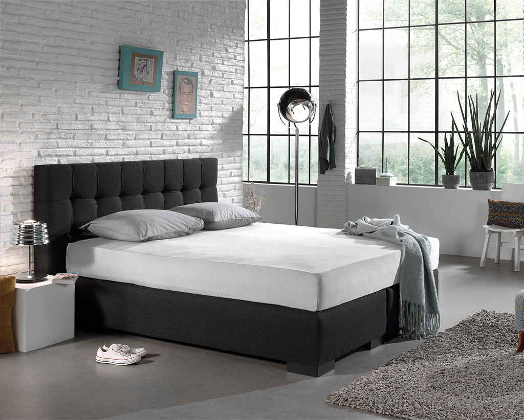 Wit hoeslaken strak om een boxspring met donkergrijs hoofdbord in een industriële slaapkamer met baksteenwand