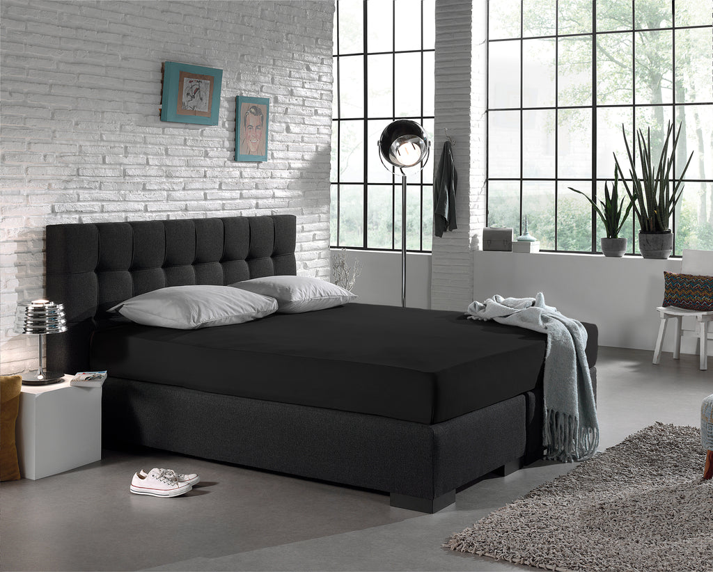 Zwart jersey hoeslaken op een boxspring met donker hoofdbord en twee grijze kussens in een industriële slaapkamer