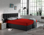 Rood jersey hoeslaken op een boxspring met grijze kussens en plaid in een slaapkamer met bakstenen muur