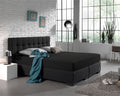 Zwart hoeslaken over een boxspring met donker gecapitonneerd hoofdbord in een lichte slaapkamer met witte bakstenen muur