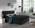 Zwart hoeslaken over een boxspring met donker gecapitonneerd hoofdbord in een lichte slaapkamer met witte bakstenen muur