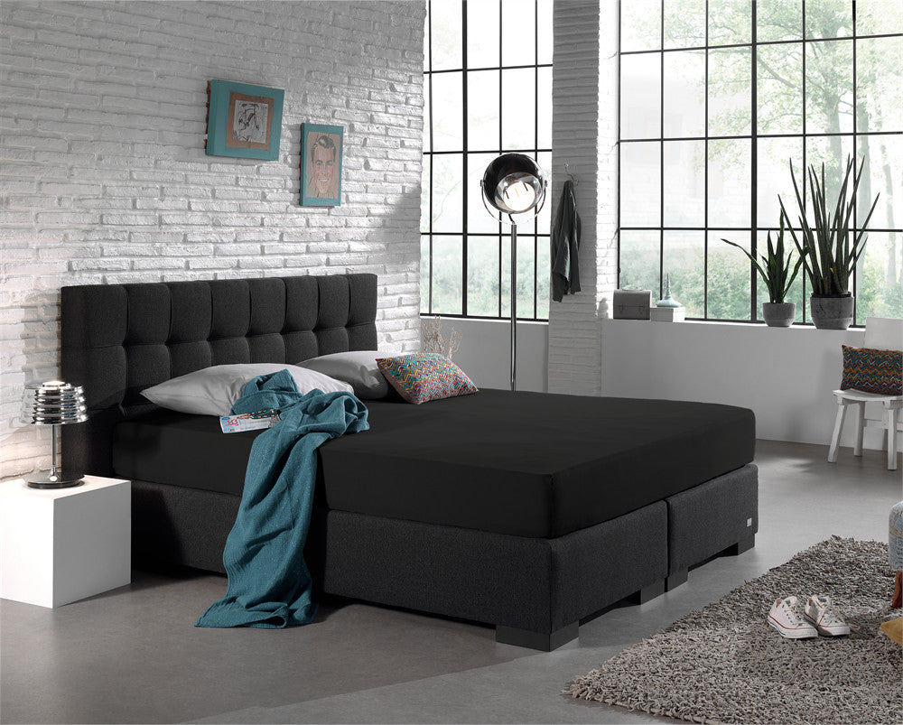 Zwart hoeslaken over een boxspring met donker gecapitonneerd hoofdbord in een lichte slaapkamer met witte bakstenen muur
