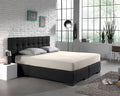 Crèmekleurig hoeslaken op een tweepersoonsbed met donkere boxspring en grijze kussens in een lichte slaapkamer