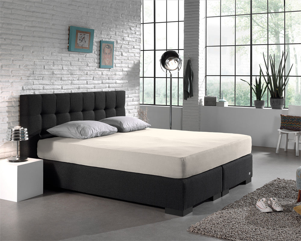 Crèmekleurig hoeslaken op een tweepersoonsbed met donkere boxspring en grijze kussens in een lichte slaapkamer