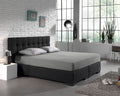 Grijs hoeslaken op een boxspring met donkergrijs hoofdbord en twee grijze kussens in een lichte slaapkamer