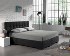 Grijs hoeslaken op een boxspring met donkergrijs hoofdbord en twee grijze kussens in een lichte slaapkamer