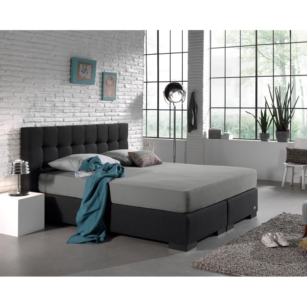 Grijs hoeslaken op een boxspring met donkergrijs hoofdbord in een slaapkamer met witte bakstenen muur