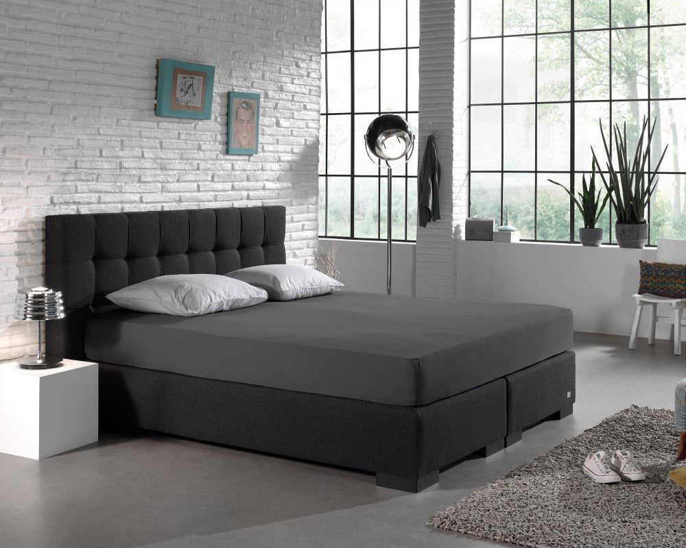 Antraciet hoeslaken op een boxspring met donker hoofdbord en twee lichtgrijze kussens in een industriële slaapkamer