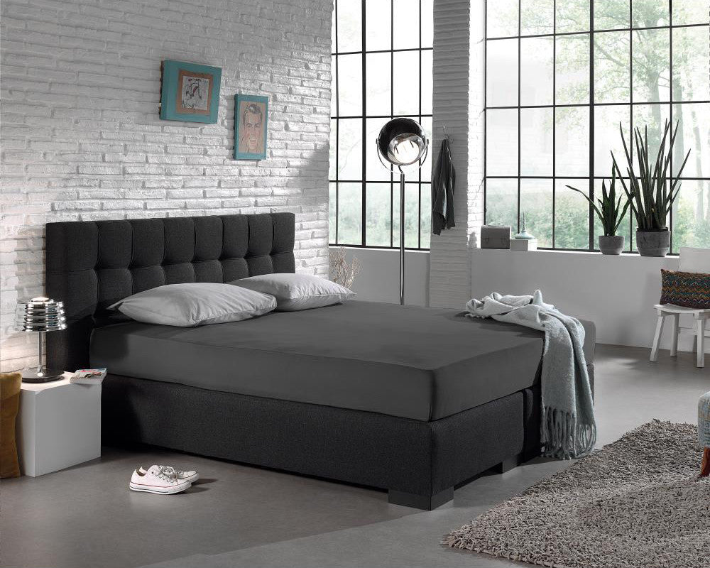 Antraciet hoeslaken op een boxspring met donker hoofdbord en twee grijze kussens in een lichte slaapkamer