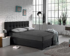 Antraciet hoeslaken op een boxspring met donker hoofdbord en twee grijze kussens in een lichte slaapkamer