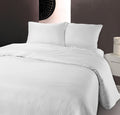 Opgemaakt bed met wit dekbedovertrek met hotelstrepen en twee bijpassende kussens, naast een wit nachtkastje