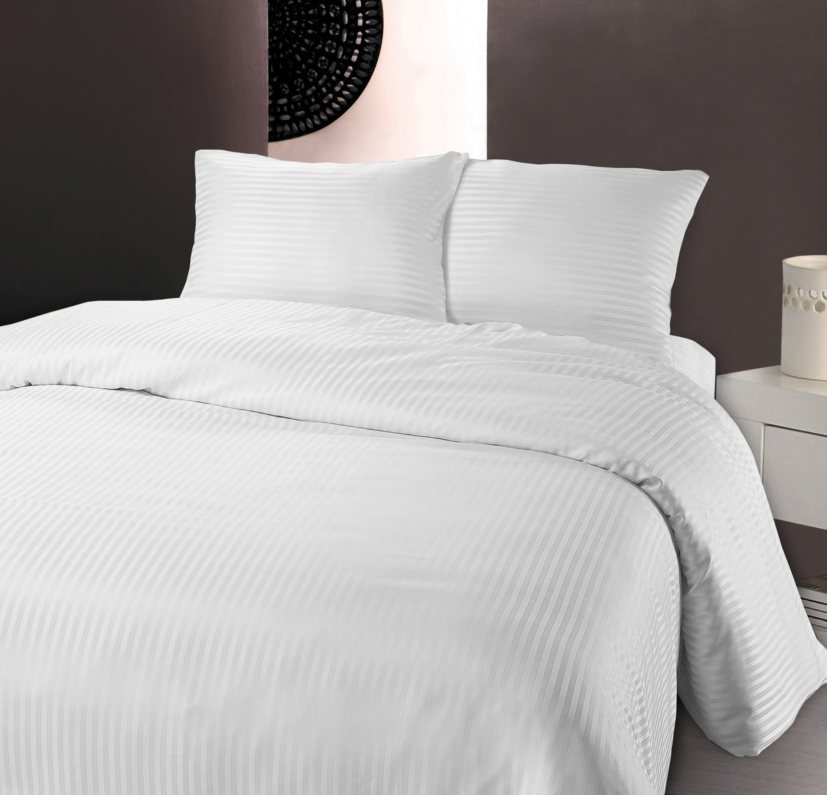 Opgemaakt bed met wit dekbedovertrek met hotelstrepen en twee bijpassende kussens, naast een wit nachtkastje