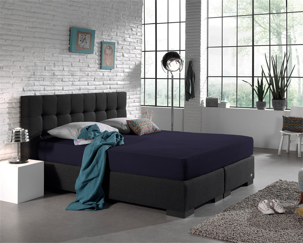 Marineblauw hoeslaken op een boxspring met donker gecapitonneerd hoofdbord in een lichte slaapkamer