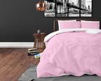 Bed met roze dekbedovertrek en roze kussenslopen met wit binnenwerk, in een donkere slaapkamer met houten vloer
