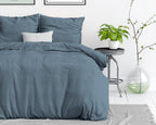 Blauw stonewashed dekbedovertrek op een opgemaakt bed met kussens, naast een bijzettafel met plant en glazen vaas