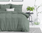 Bed opgemaakt met groen stonewashed dekbedovertrek en kussens, naast een bijzettafel met plant en glazen vaas