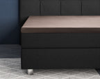 Taupe satijnen hoeslaken over een topper op een donkergrijs boxspringbed met gecapitonneerd hoofdbord