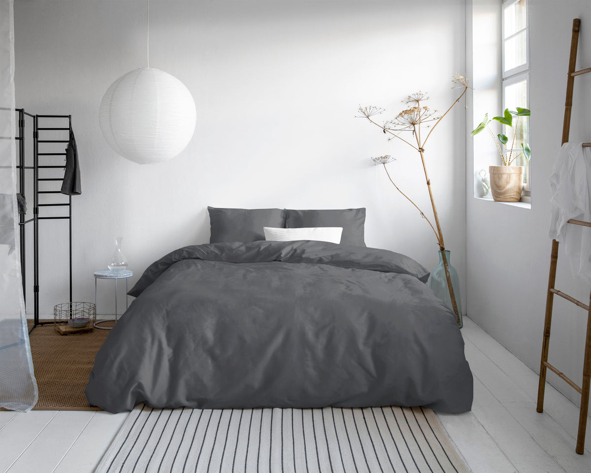 Bed opgemaakt met antraciet dekbedovertrek en kussenslopen in een lichte slaapkamer met witte hanglamp