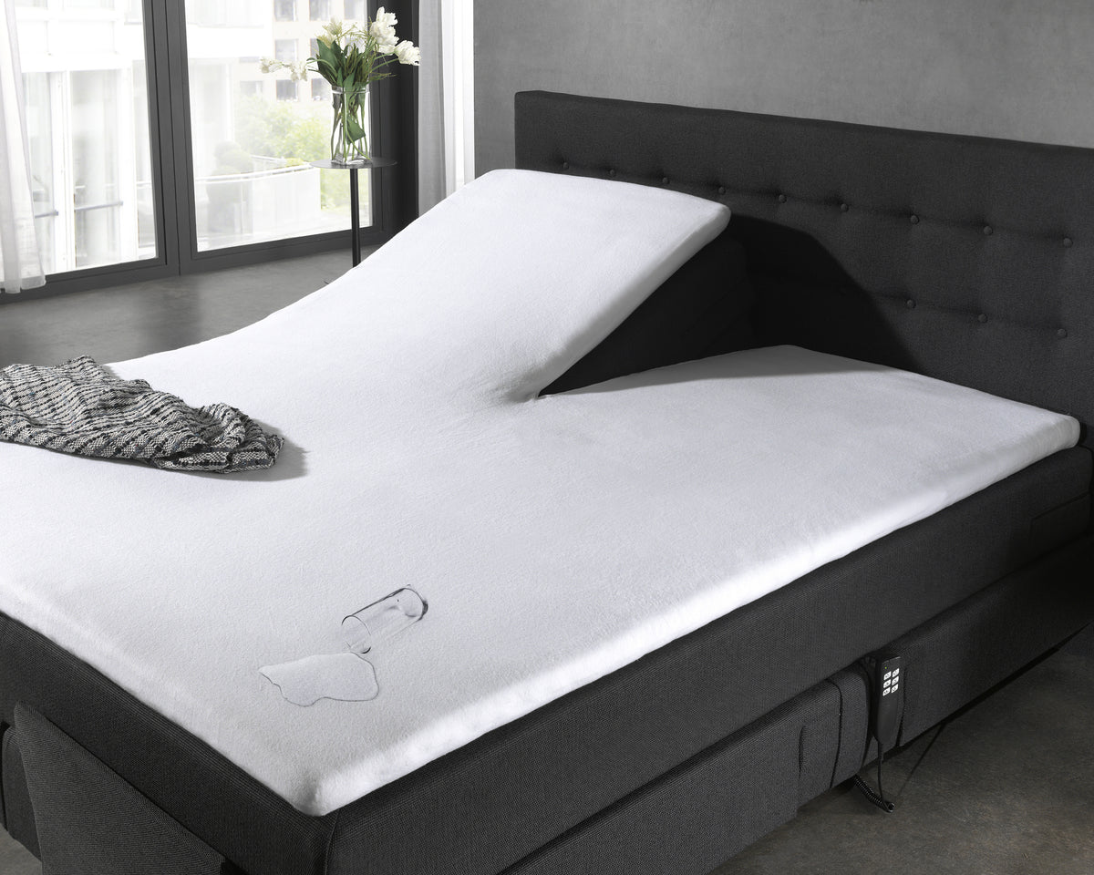Wit waterdicht hoeslaken op een split-topper van een verstelbaar bed met omgevallen glas en gemorst water