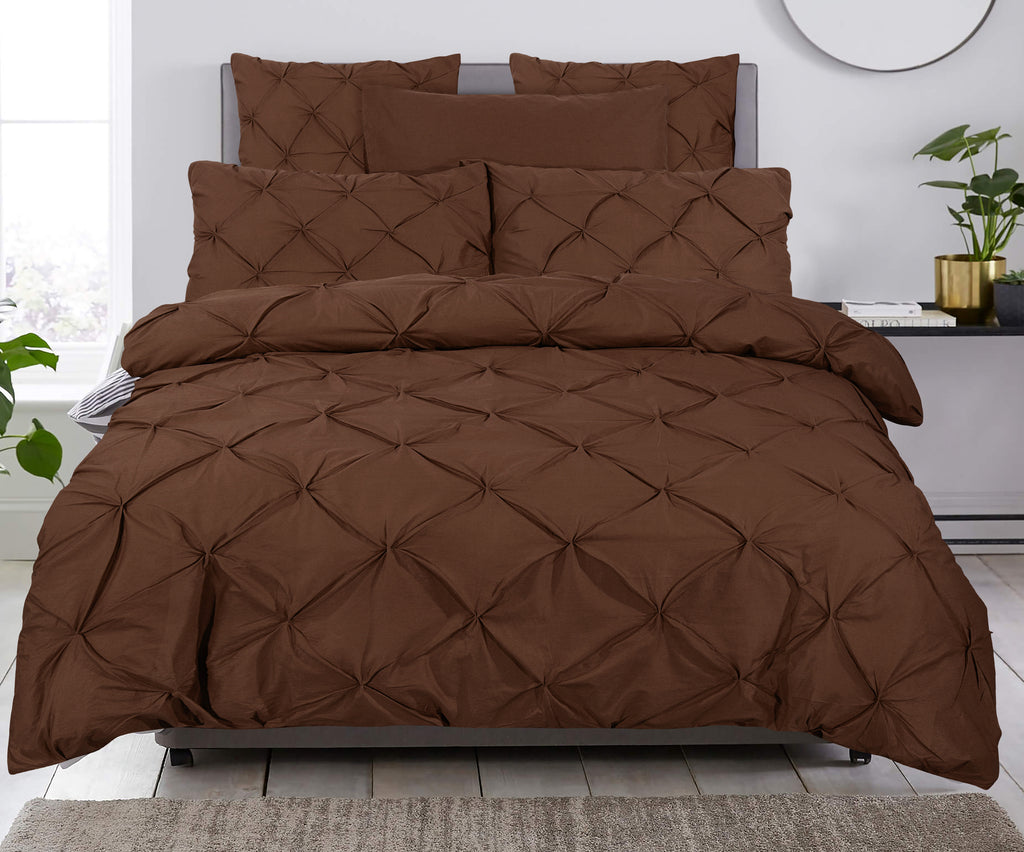 Bed met bruin pintuck dekbedovertrek en bijpassende kussens tegen een lichte muur, met plant en schaal ernaast