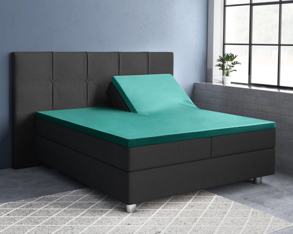 Groen satijnen splittopper-hoeslaken op een boxspring met grijs hoofdbord, één helft van het hoofdeinde omhoog