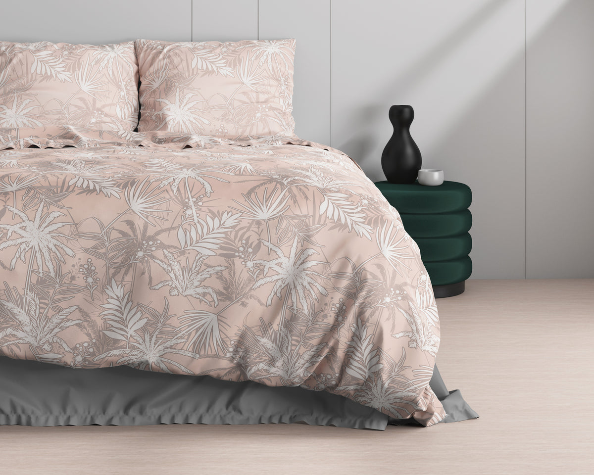 Roze dekbedovertrek met witte en taupe palmbladeren op een bed met twee kussens, naast een groene poef