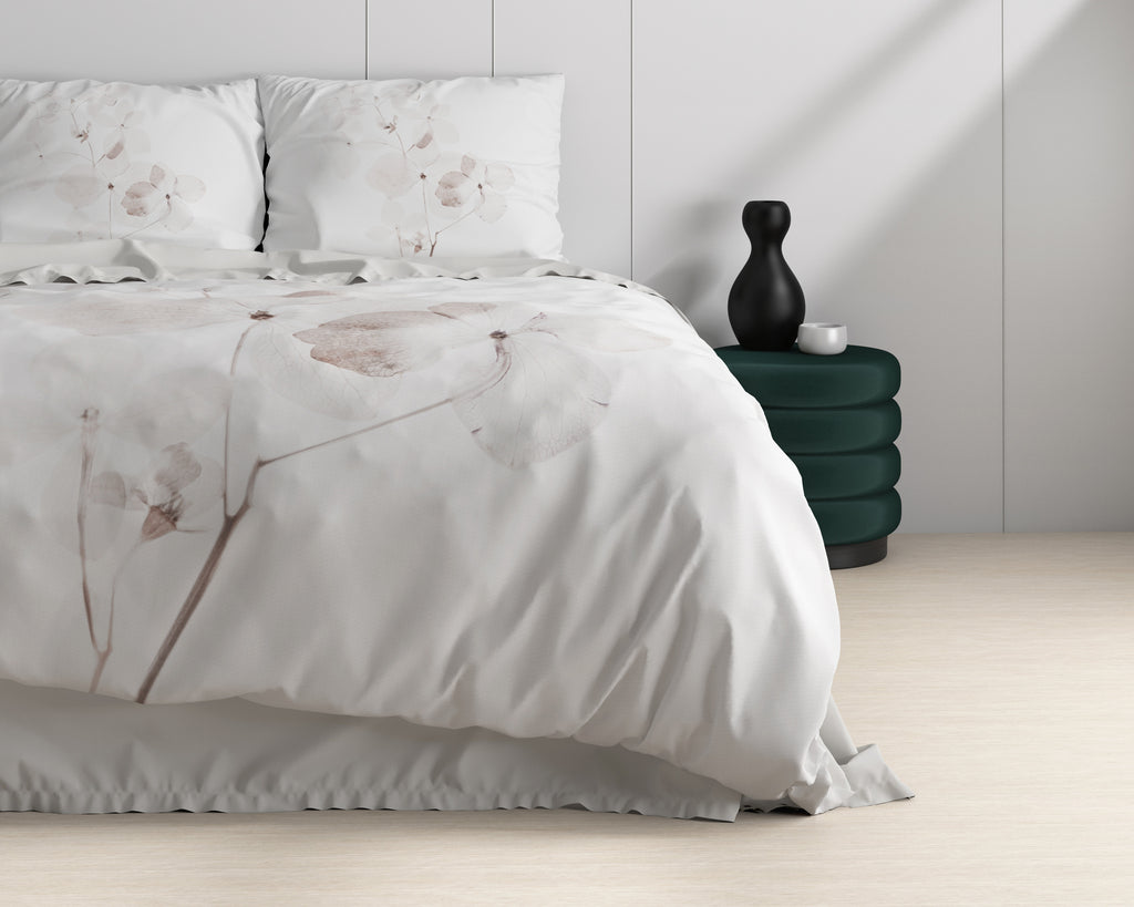 Wit dekbedovertrek met subtiel bloemendessin op een bed, naast een groen poefje met zwarte vaas