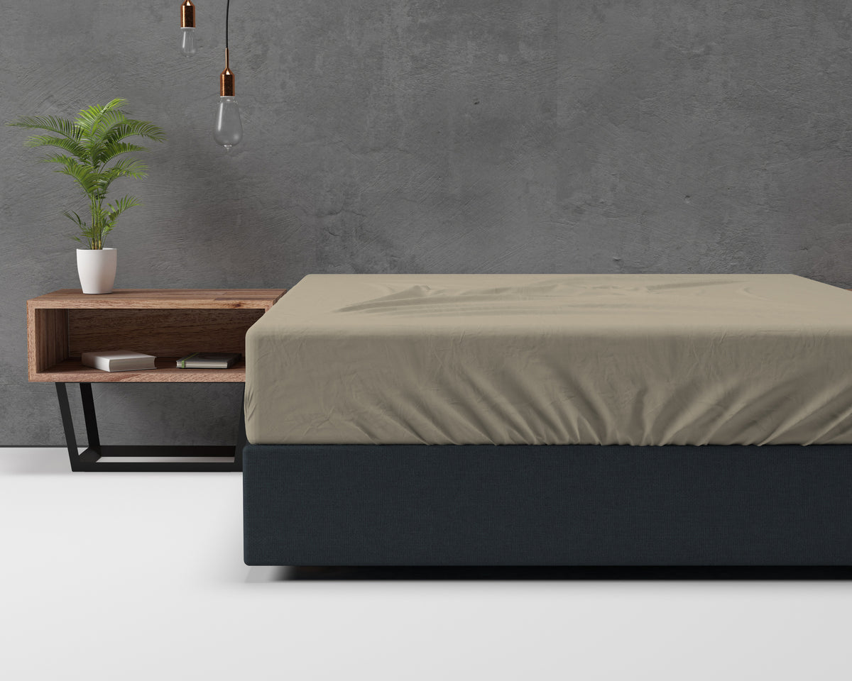 Taupe hoeslaken op een bed met donkere bekleding, naast een houten nachtkastje en een plant tegen een betonnen wand