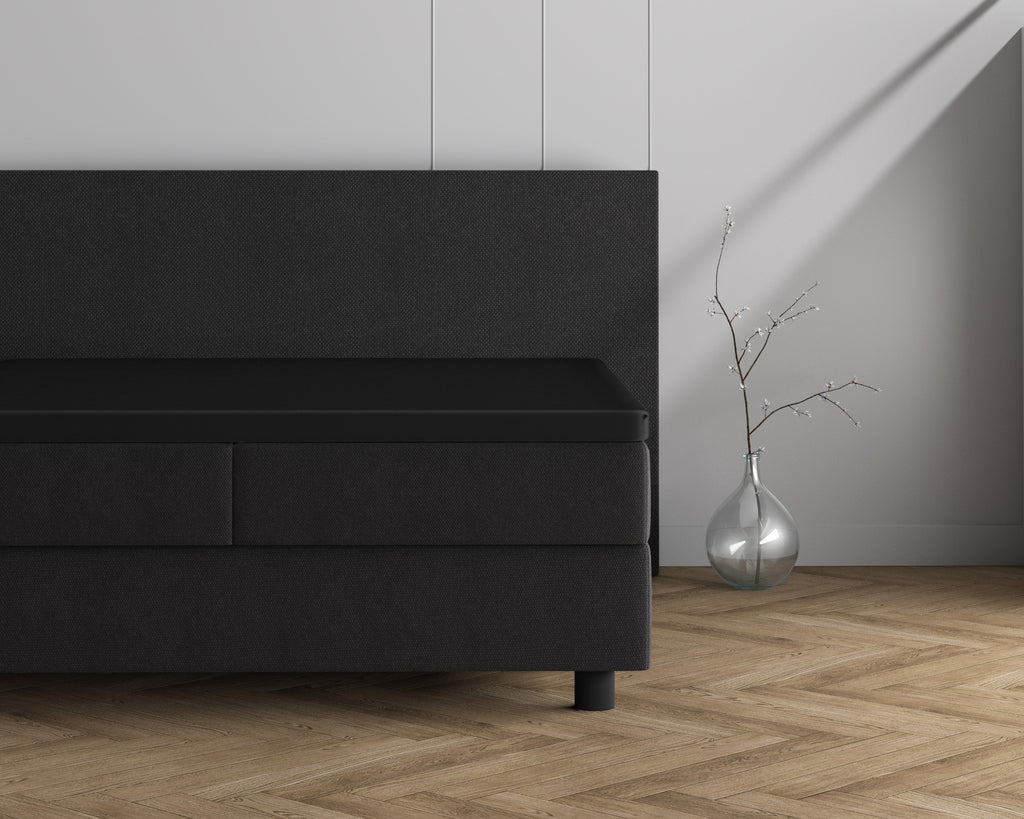 Zwarte topper met hoeslaken op een donkergrijs boxspringbed met hoofdbord, naast een glazen vaas met tak