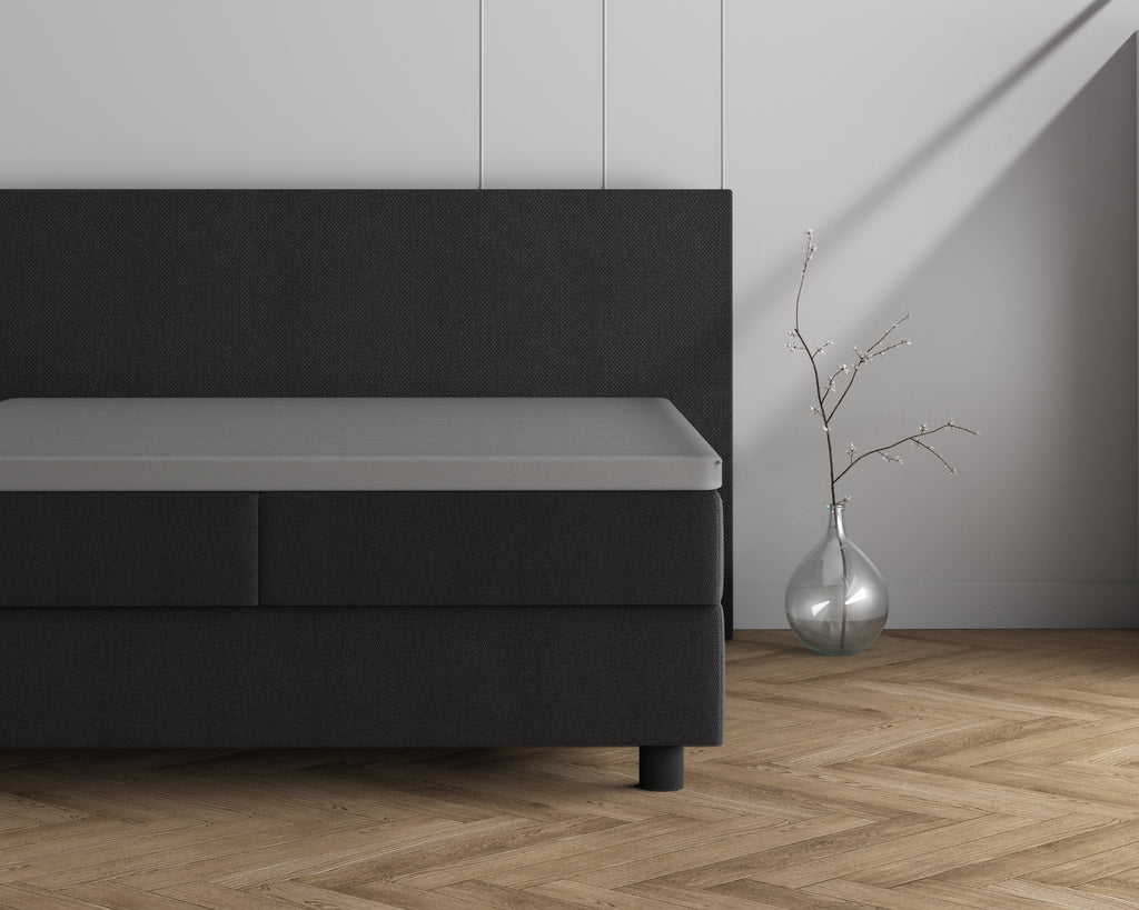 Grijs topper hoeslaken op een donkergrijs boxspringbed naast een glazen vaas met takje op een visgraatvloer