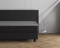 Grijs topper hoeslaken op een donkergrijs boxspringbed naast een glazen vaas met takje op een visgraatvloer
