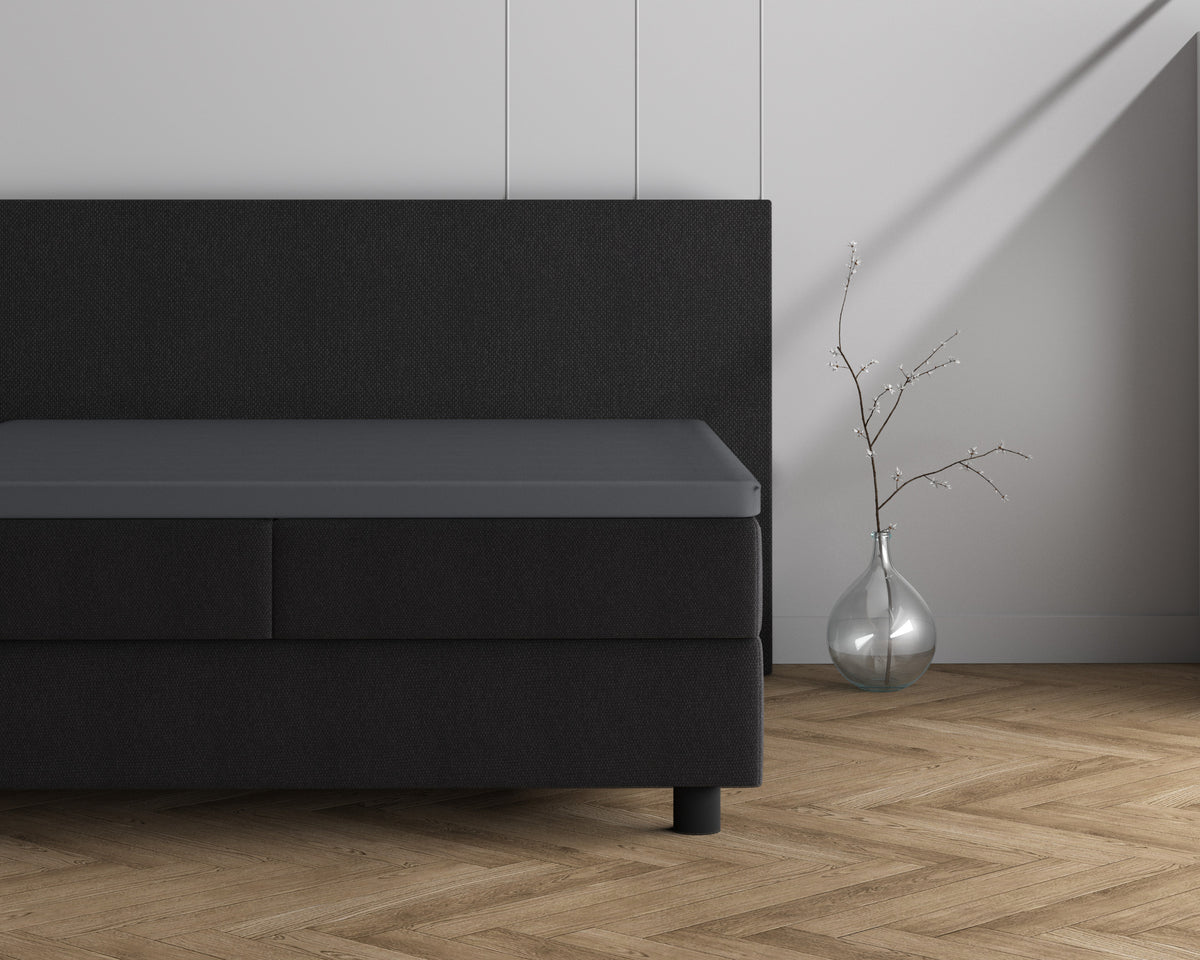 Antraciet hoeslaken over een topper op een donkere boxspring, naast een glazen vaas met een takje