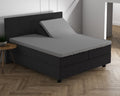 Grijs split-topper hoeslaken op een boxspring met donker hoofdbord, één helft van de topper omhoog gezet