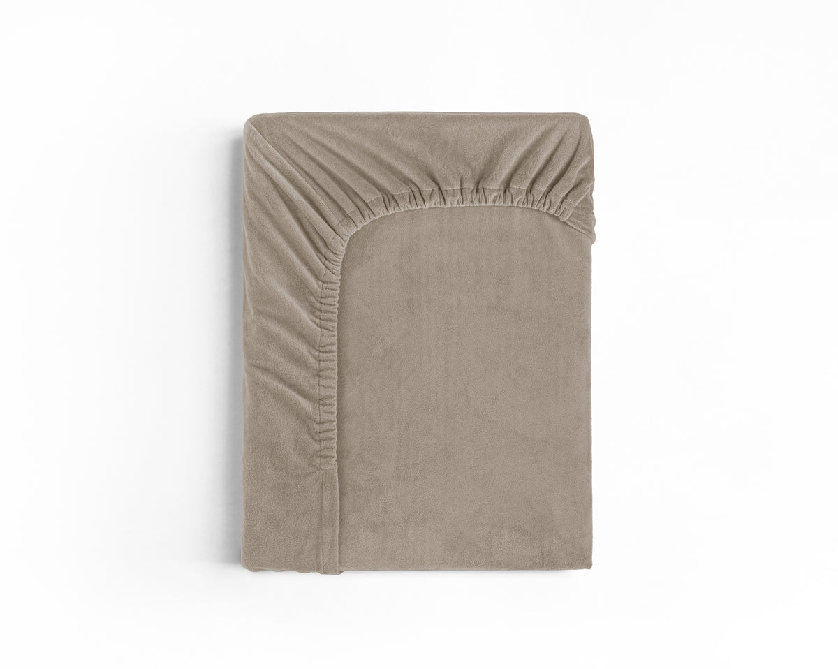 Opgevouwen taupe hoeslaken van zachte velvet stof met elastische rand, bovenaanzicht op witte achtergrond