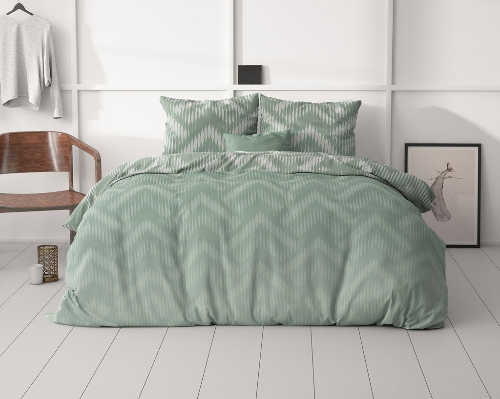 Bed met groen dekbedovertrek met zigzagpatroon en kussens, in een lichte slaapkamer met houten stoel