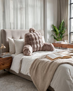 Taupe bedsteunkussen van nepbont op een bed met wit beddengoed, beige plaid en een opengeslagen boek