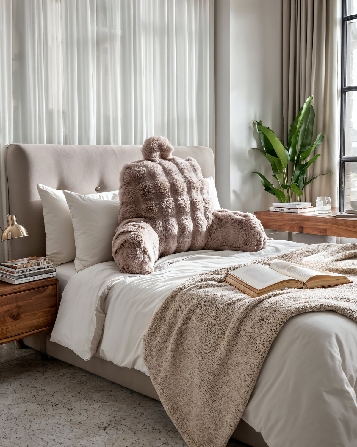 Taupe bedsteunkussen van nepbont op een bed met wit beddengoed, beige plaid en een opengeslagen boek
