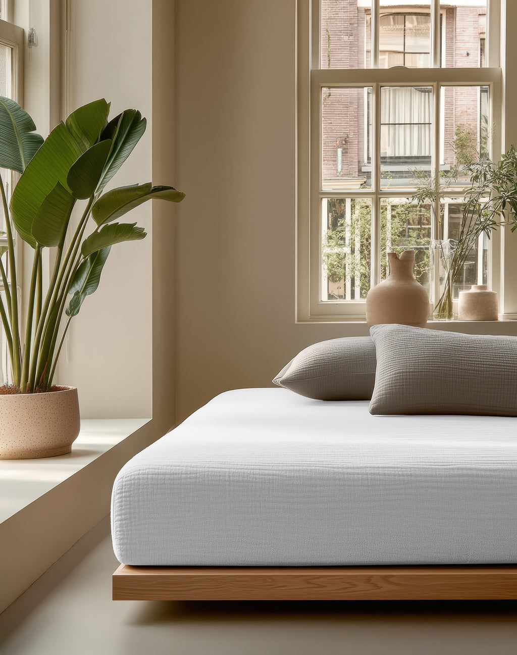 Wit mousseline hoeslaken om een matras op een houten bedframe, met grijze kussens in een lichte slaapkamer