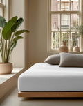 Wit mousseline hoeslaken om een matras op een houten bedframe, met grijze kussens in een lichte slaapkamer