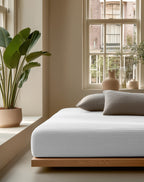 Wit mousseline hoeslaken om een matras op een houten bedframe, met grijze kussens in een lichte slaapkamer