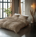 Bed met beige-bruin dekbedovertrek met bladmotief en witte kussens in een slaapkamer met hoge ramen en hanglamp