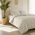 Beige gewatteerd dekbed met beige kussens op een bed naast een palm in rieten mand in een lichte slaapkamer
