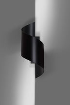 Zwarte spiraalvormige wandlamp aan een grijze muur met zacht licht dat langs de wand valt