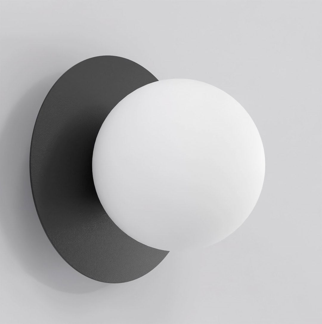Zwarte wandlamp met ronde achterplaat en een witte glazen bol aan een lichtgrijze muur