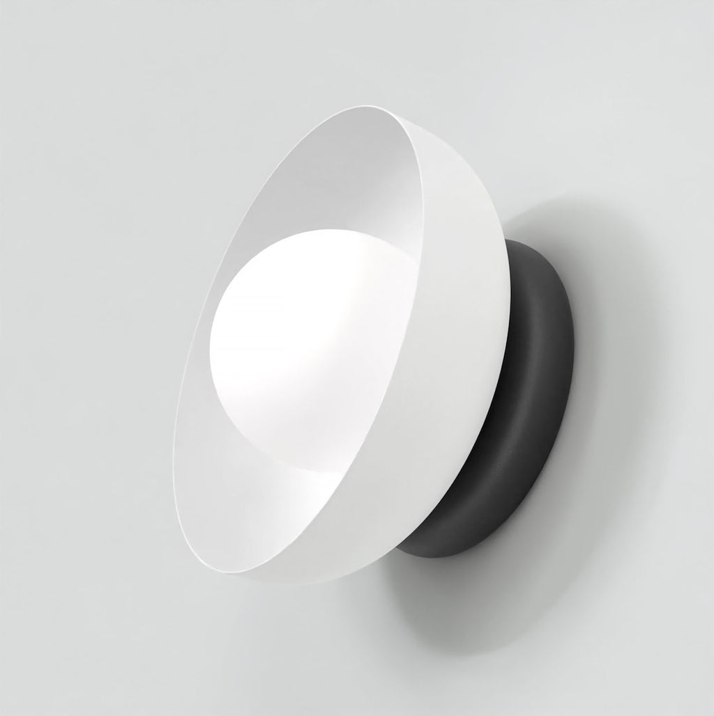 Witte wandlamp met schuin geplaatste kap, lichtgevend bolletje en zwarte ronde basis tegen een lichte wand